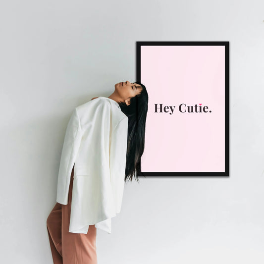 Woman leaning against a wall with a framed poster displaying 'Hey Cutie.' Framiqa wall art for salons and beauty parlours